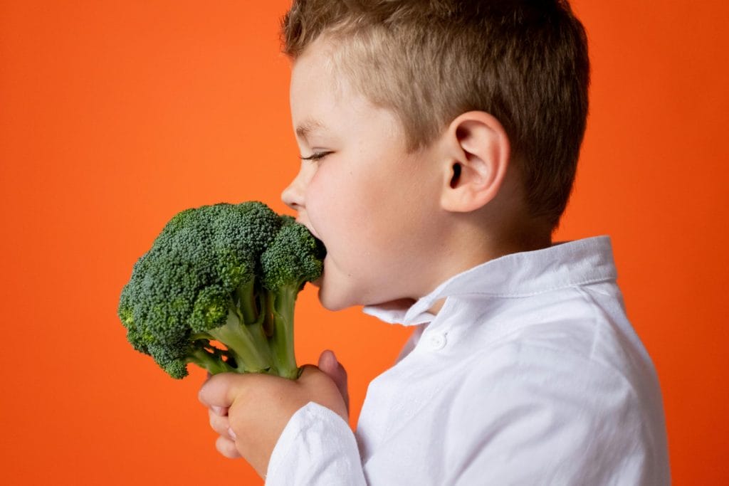 A young child biting into fresh broccoli against an orange background.