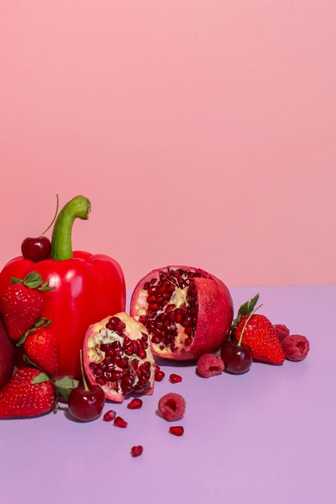 A colorful still life of red fruits and vegetables against pink and purple backgrounds.