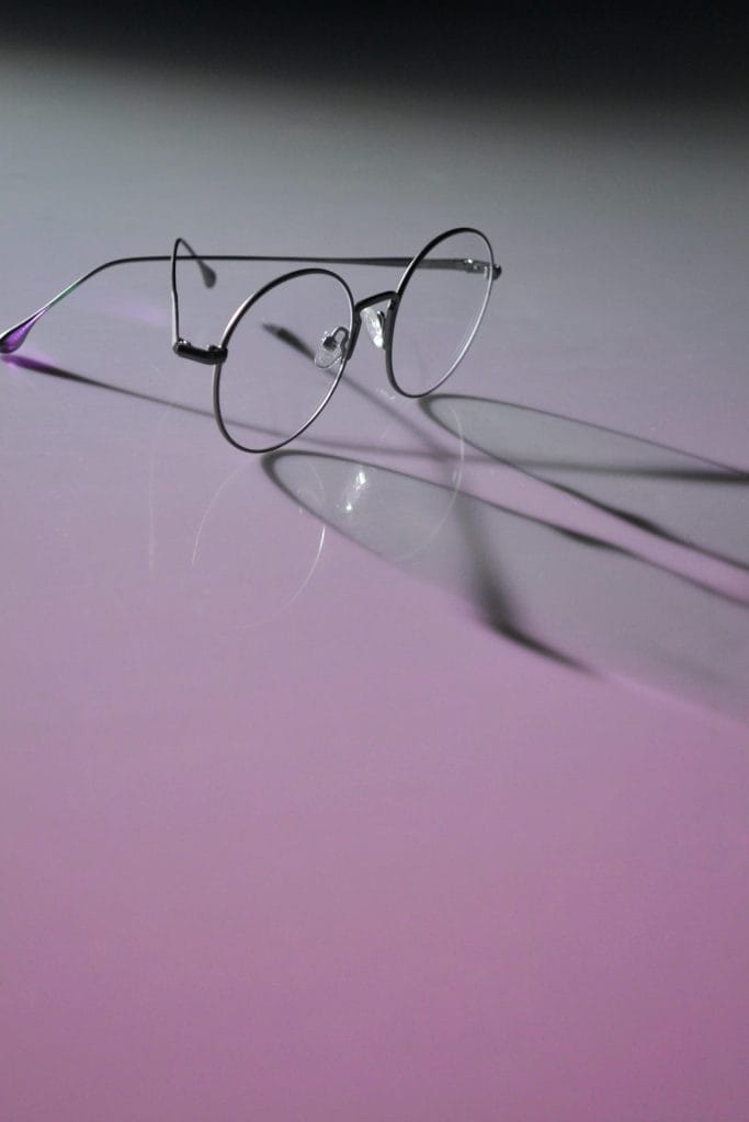 Round eyeglasses with metal frames casting shadows on a neon purple surface, emphasizing modern eyewear style.