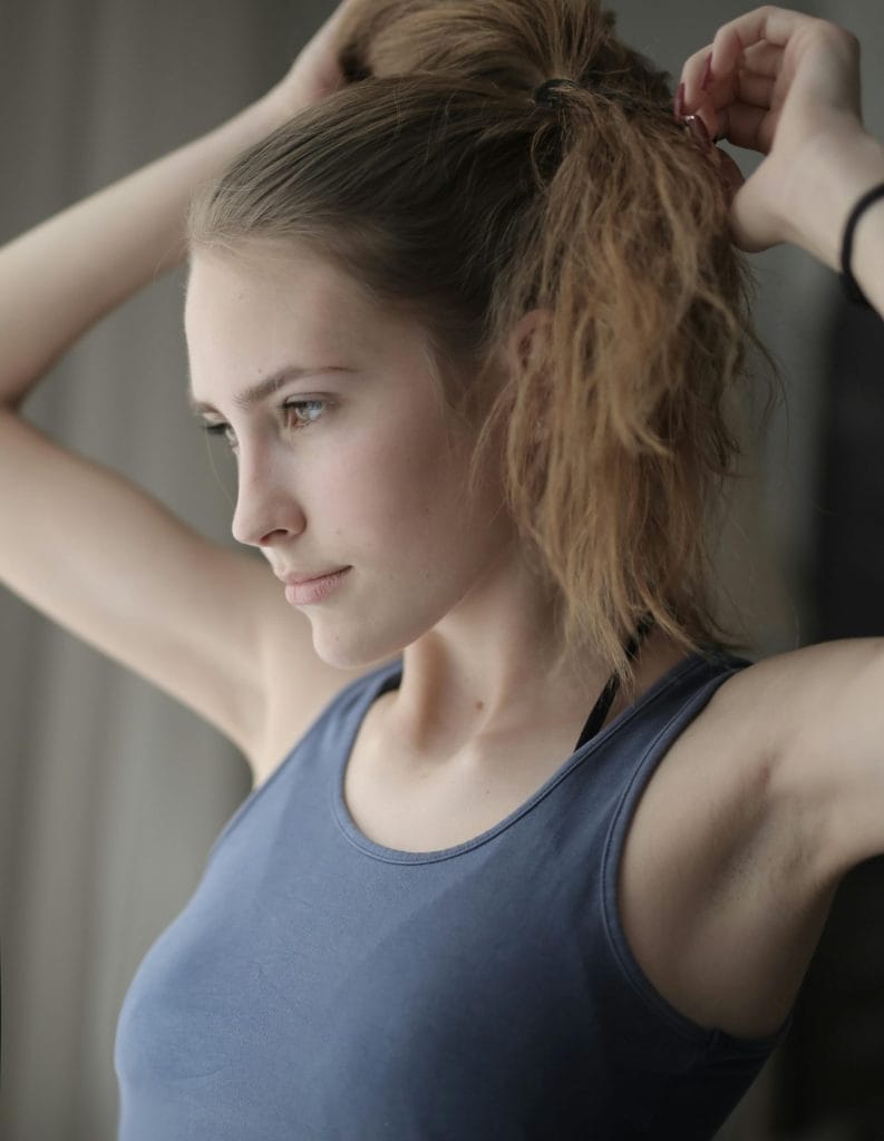 Side view of young female with natural beauty in gray t shirt with brown hair making ponytail and looking forward seriously