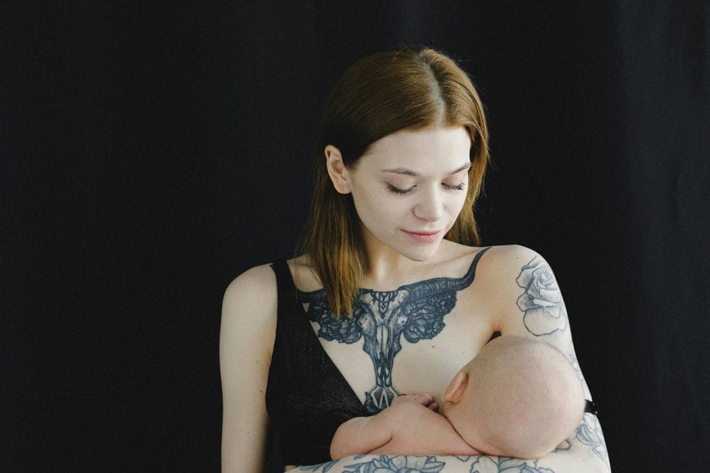 A mother with tattoos lovingly breastfeeding her infant against a dark backdrop.