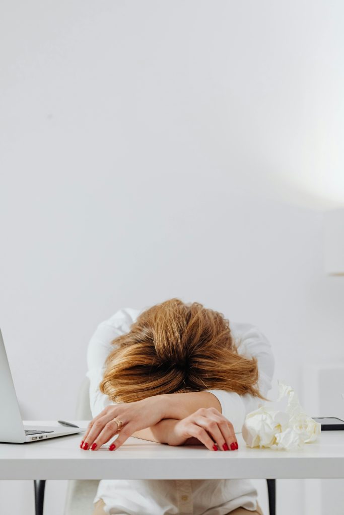 A tired woman with red nails resting her head on a white desk in a bright office setting.