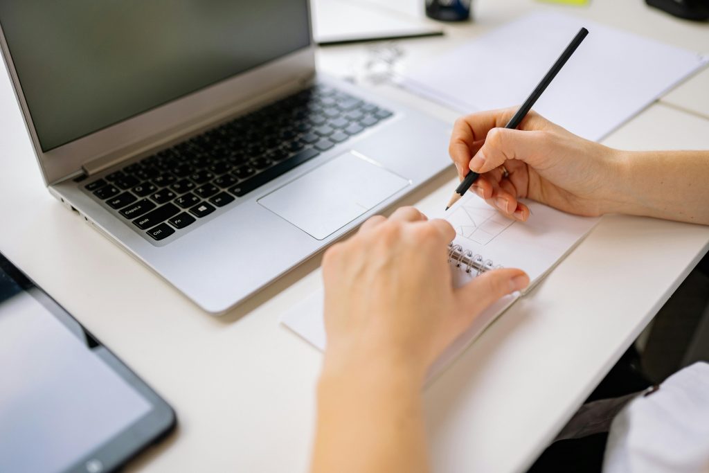 Hands sketching in a notebook beside a laptop on a desk, capturing creativity.