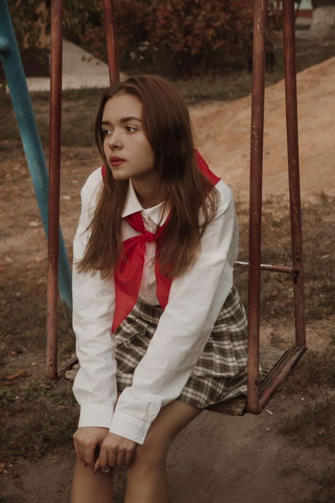 Thoughtful teenager with brown hair on a swing wearing a red scarf and plaid skirt outdoors.
