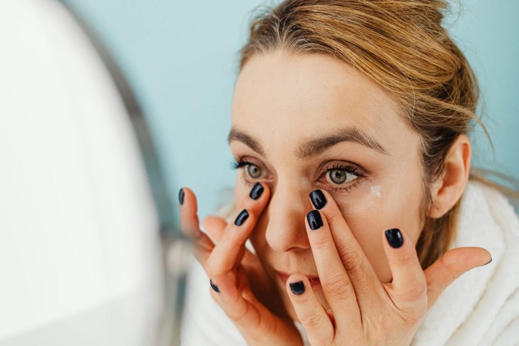 Close-up of a woman applying facial cream in front of a mirror with blue background.