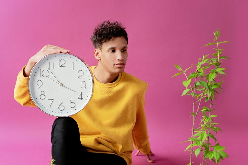 A young man sitting with a clock and green plant against a pink studio backdrop.